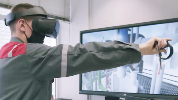 Azubi testet Chemikant VR, auf einem Fernseher im Hintergrund sieht man, wie er an den virtuellen Ventilen dreht.